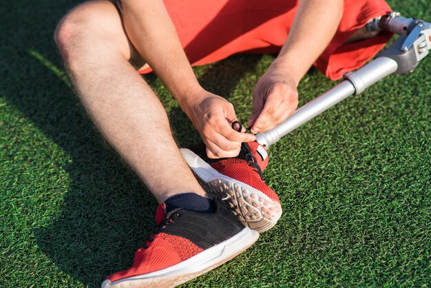 Close up of a caucasian guy with a prosthetic leg sitting on\
the field in the stadium and tying a lace on a sneaker.