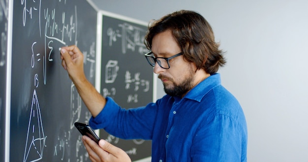 Close up of Caucasian male teacher in glasses writing formulas and math laws on blackboard and looking at smartphone. Educative mathematics lection. Man lecturer using mobile phone as cribe.