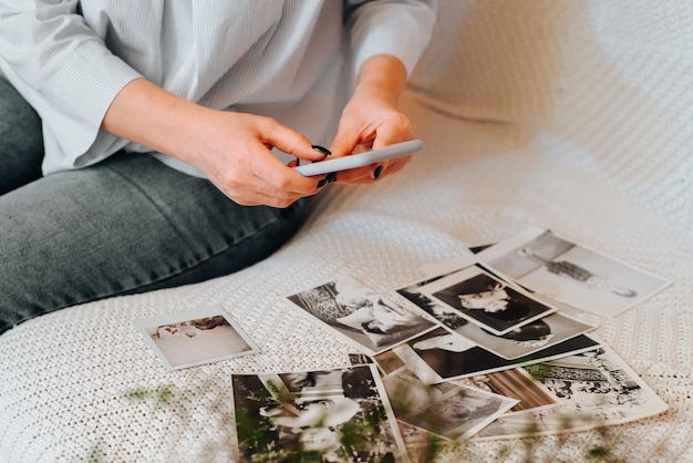 Photo close-up of caucasian woman photographing old black-and-white photos from family album on smartphone camera while sitting on sofa, indoors. selective focus on female hands holding mobile phone