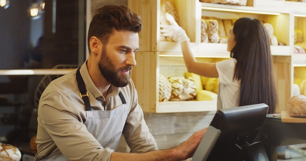 Close up of Caucasian young male and female bread vendors selling baguettes to the buyer at the counter in the bakery shop. Indoors.