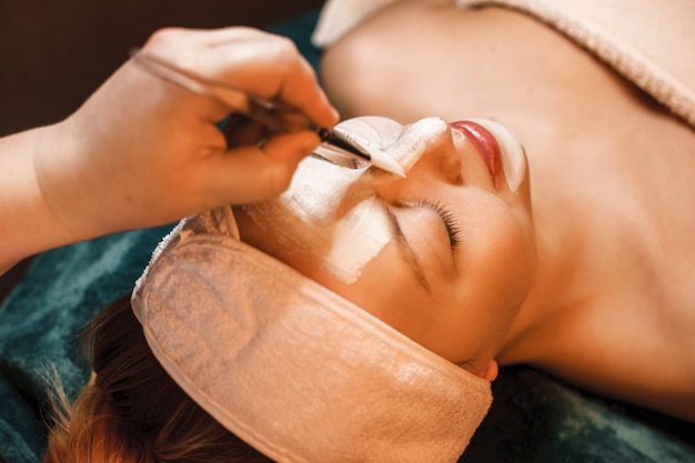 Close up of a charming woman leaning on spa bed with closed eyes doing a white skin cleansing mask .