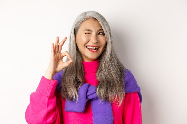 Close-up of cheerful asian grandmother in fashionable pink sweater, winking and showing okay sign, recommending promotion, standing over white background