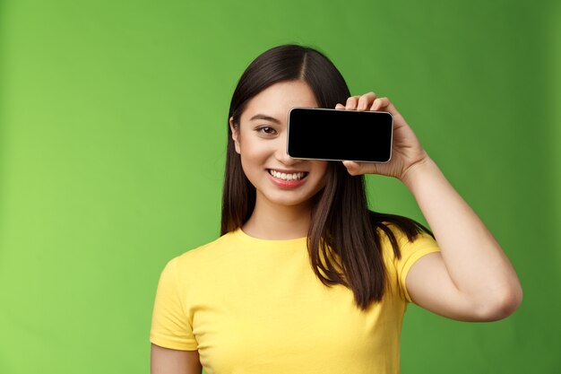 Крупным планом веселая оптимистичная азиатская женщина продвигает приложение для смартфона, держит телефон рядом с глазом, камера удовлетворена, широко улыбается, представляет игру, крутое новое приложение, стоит на зеленом фоне