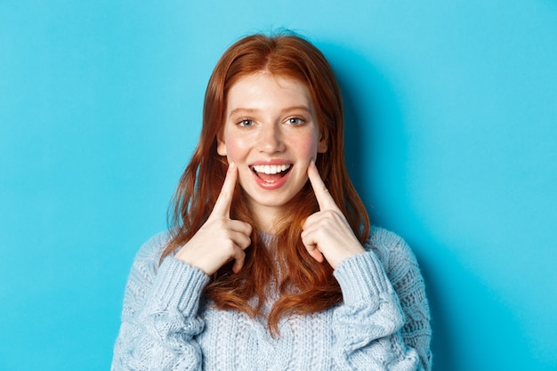 Close-up of cheerful teenage girl with red hair and freckles, poking cheeks, showing dimples and smiling with white teeth, standing over blue background.