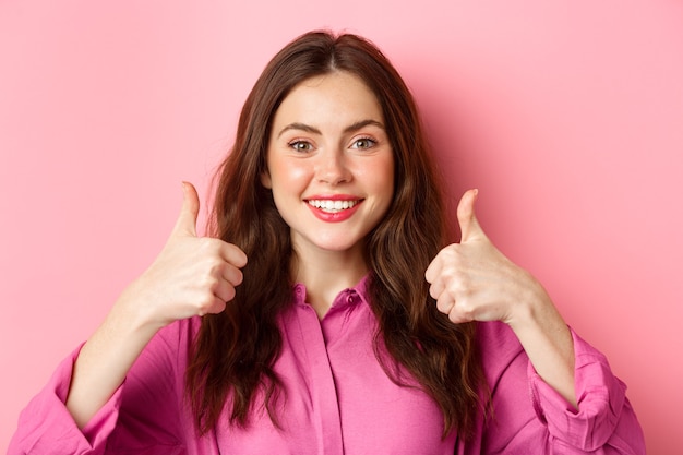 Photo close up of cheerful young lady fully approve your choice, praising something good, showing thumbs up and smiling pleased, pink wall.