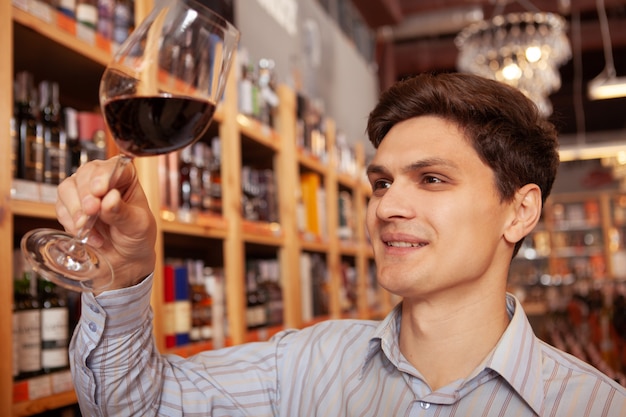 Chiuda in su di un vino allegro dell'assaggio del giovane alla cantina. messa a fuoco selettiva sul bicchiere di vino rosso nella mano di un uomo felice