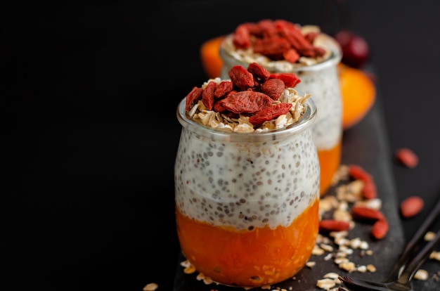 Close up of chia seed pudding with goji berries, smashed fresh apricot and oat meals on aslate board on black