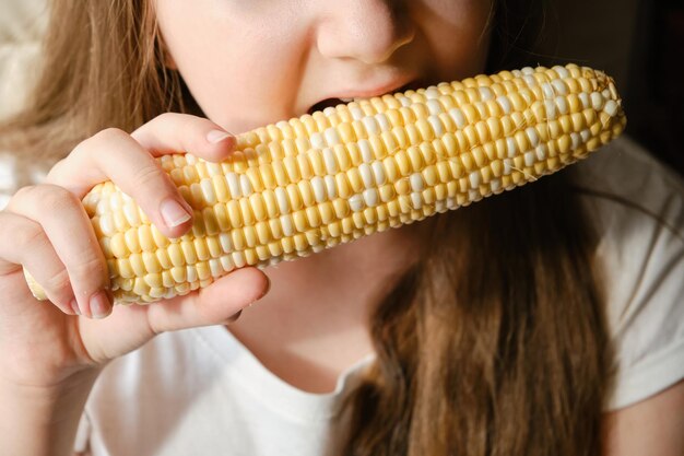 Close up child girl biting big vegetable corn at home fresh vegetarian food vitamins healthy nutrition without GMOs indoor unrecognizable face