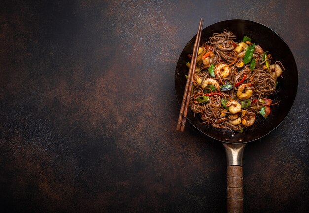 Close-up of Chinese soba stir-fry noodles with shrimps, vegetables in old rustic wok pan served on concrete background, close up, top view. Traditional asian/thai dish, space for text