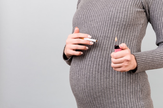 Close up of a cigarette and a lighter in pregnant woman's hands at gray background. Unhealthy habit during pregnancy. Smoking abuse.