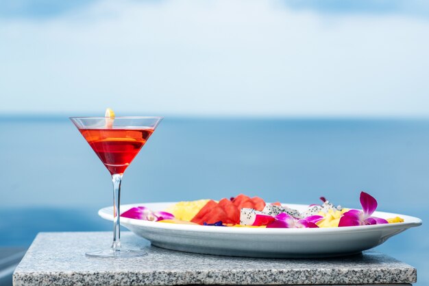 Close-up the cocktail is cosmopolitan with a slice of lemon and next to it is a plate with exotic fruits