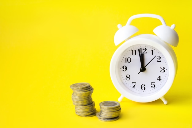 黄色の背景の目覚まし時計によるコインのクローズアップ。節約の概念、時間の財務の概念。