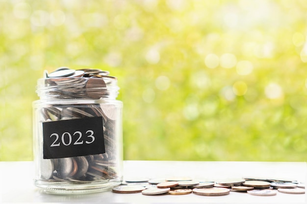 Close up of coins in glass jar on white table over green bokeh background Collect money for future saving and investment in 2023 concept Closeup copy space