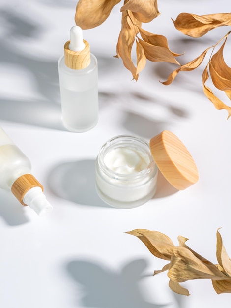 Close up of collagen cream in glass jar serum in white matte glass bottle and dry ruskus leaves on white background Set for skin and body care beauty products