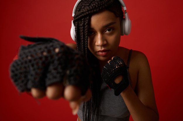 Close-up of concentrated young African athletic woman boxer in headphones and red boxing gloves striking a straight punch. Contact martial art concept isolated on colored background with copy space