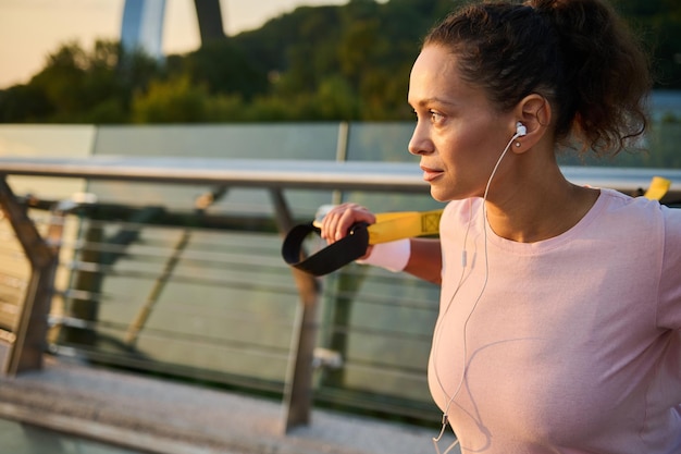 Close-up of confident determined middle aged african american\
sportswoman, female athlete exercising outdoor with suspension\
straps, doing body weight intensive training on the city bridge at\
sunrise