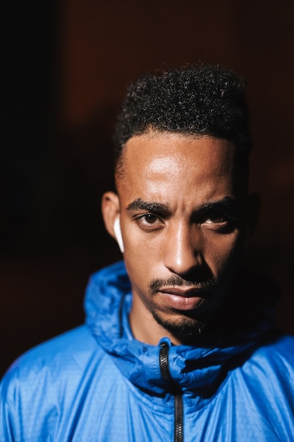 Close up of a confident fit young african sports man wearing windbreaker exercising outdoors at night time, wearing wireless earphones