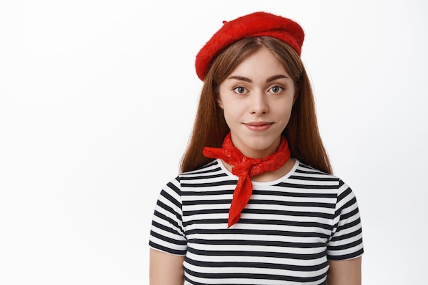 Foto primo piano di una giovane donna sicura di sé in berretto francese e sciarpa rossa intorno al collo, sorridente e dall'aspetto determinato davanti, muro bianco