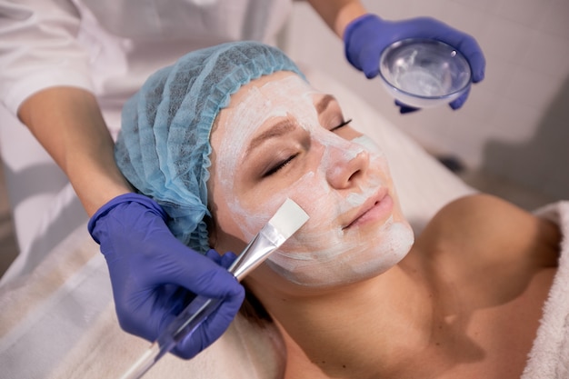 Close up of cosmetologist's hands in blue gloves applying a medical mask to the patient's face with a brush