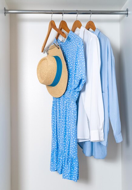 Photo close up cotton dress and blouses with straw hat hanging on wooden racks in modern white wardrobe