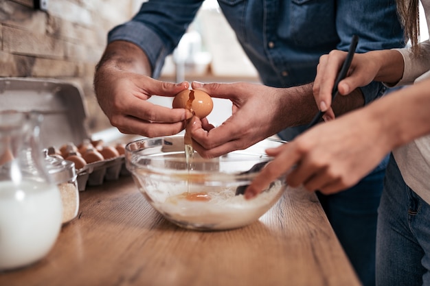 Photo close-up of couple cooking and breaking eggs into bowl in the kitchen at home.