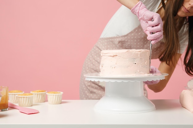 close up cropped chef cook confectioner or baker in white t-shirt cooking at table isolated on pink pastel background in studio. cream application cake making process. mock up copy space food concept.