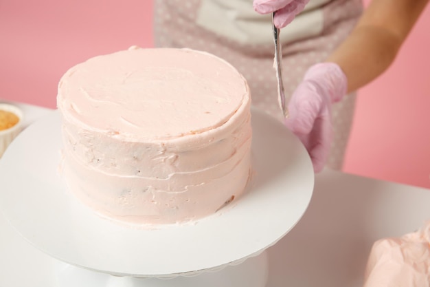 close up cropped chef cook confectioner or baker in white t-shirt cooking at table isolated on pink pastel background in studio. cream application cake making process. mock up copy space food concept.