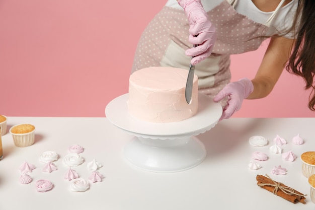 Close up cropped chef cook confectioner or baker in white t-shirt cooking at table isolated on pink pastel background in studio. Cream application cake making process. Mock up copy space food concept.