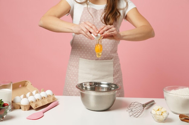 Close up cropped female chef cook confectioner or baker in apron white t-shirt cooking cake or cupcake at table hold eggs isolated on pink pastel background in studio. Mock up copy space food concept.