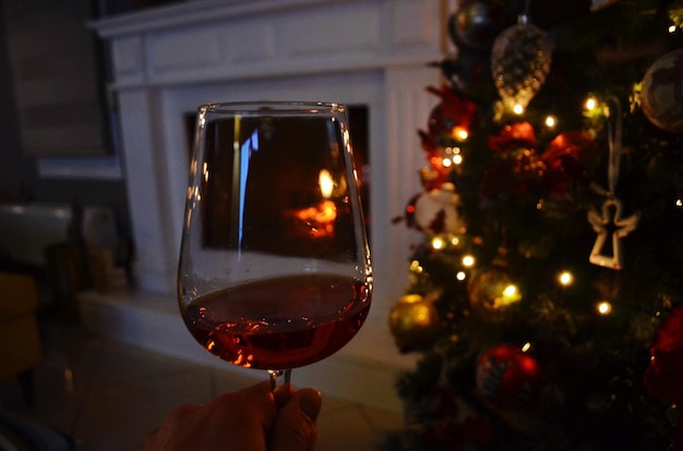 Close-up di una mano tagliata che tiene il vino rosso in un bicchiere vicino all'albero di natale illuminato