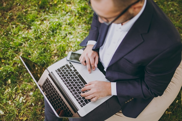 Close up cropped of hands on the keyboard. Businessman in classic suit, glasses. Man sit on soft pouf, work on laptop pc computer in city park on green lawn outdoors. Mobile Office concept. Top view.