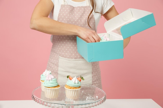 Close up cropped housewife female chef cook confectioner or\
baker in apron white t-shirt packaging cake cupcake at table\
isolated on pink pastel background in studio. mock up copy space\
food concept.