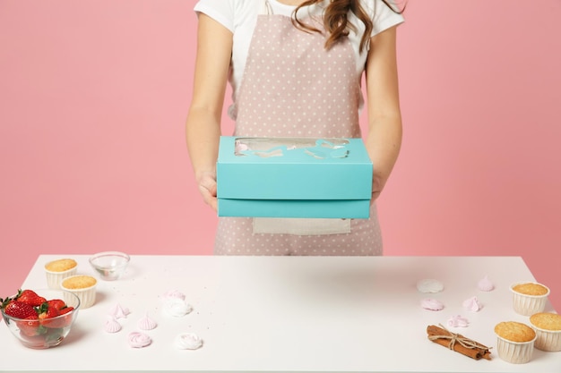 Close up cropped housewife female chef cook confectioner or\
baker in apron white t-shirt packaging cake cupcake at table\
isolated on pink pastel background in studio. mock up copy space\
food concept.