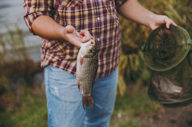 Фото Крупным планом обрезанный портрет мужчина в клетчатой рубашке держит в руках зеленую рыболовную сетку и рыбу, которую он поймал на берегу озера возле кустарников и тростников. образ жизни, отдых, концепция досуга рыбака