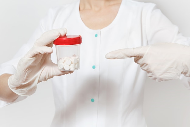 Close up cropped portrait of young doctor woman isolated on white background. Female doctor in medical gown, gloves pointing on bottle with pills. Healthcare personnel, medicine concept. Copy space.