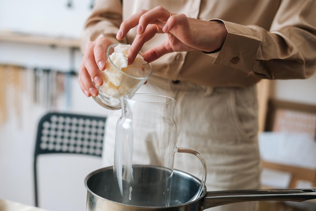 Close-up cropped shot of female artisan adding white dry soy wax in glass jar into pot of boiling water for creating candle building mixture. Process of making handmade natural candle at workshop.