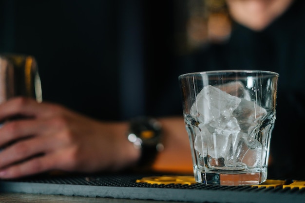 Крупный план неузнаваемого мужчины-бармена, стоящего за барной стойкой, со стаканом, наполненным кубиками льда на переднем плане. Бармен делает коктейль со льдом в современном баре с темным интерьером.
