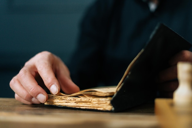 Close-up cropped shot of unrecognizable senior aged male open\
old book and turning pages sitting at table. closeup of mature man\
start reading novel and flip through pages of paper book.