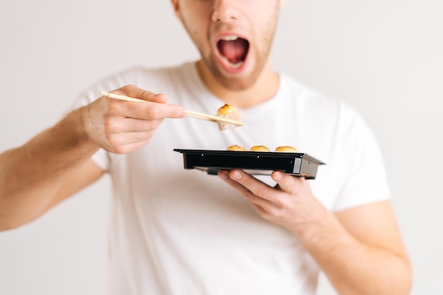 Крупный план молодого человека, наслаждающегося поеданием свежих вкусных суши-роллов с палочками для еды на белом изолированном фоне. Студийный снимок счастливого кавказца, который ест традиционную японскую еду.
