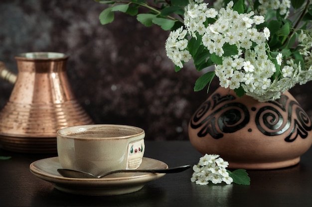Close-up of a cup of coffee and a branch of blooming spirea in a ceramic vase on a dark surface