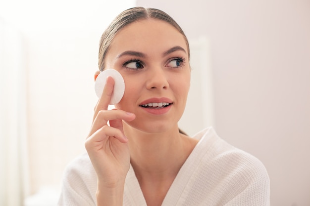 Close up of the curious young lady putting a cotton pad on her cheek and smiling while looking into the distance with interest