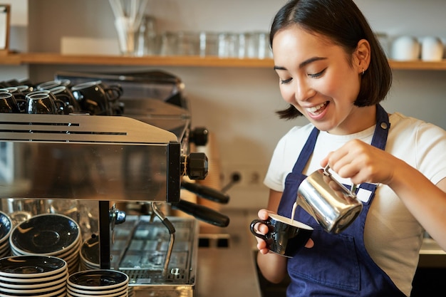 Close up of cute asian barista girl making cappuccino doing latte art in cup with steamed milk stand