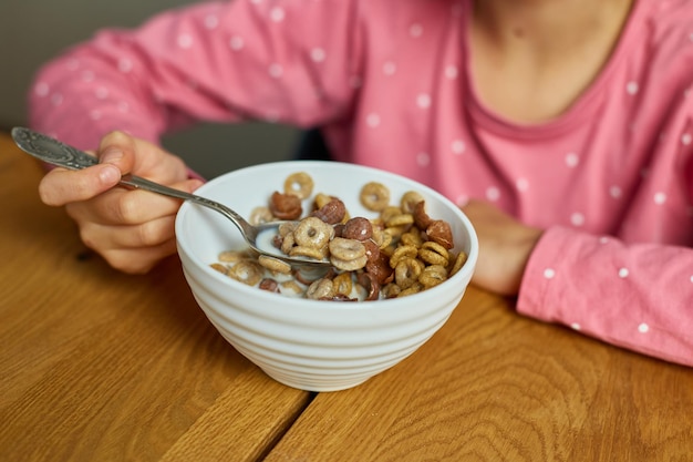 クローズアップかわいい女の子は、食欲をそそる朝の朝食のためにミルクと一緒にシリアルを食べることを楽しんでいます、どのようにおいしい健康的な食べ物、家で朝食をとっている美しい子供のゆっくりとした動き。