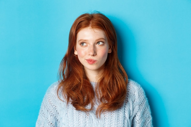 Close-up of cute redhead girl thinking and looking left, have an idea, standing against blue background