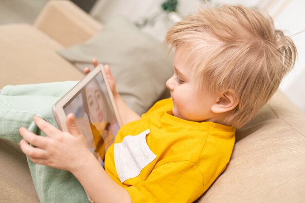 금발 머리가 소파에 앉아 태블릿을 사용하여 온라인으로 엄마와 채팅하는 동안 귀여운 아들의 근접