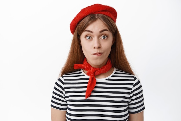 Close up of cute young woman in french beret and scarf bites lips from temptation want something looks excited and hopeful standing over white background