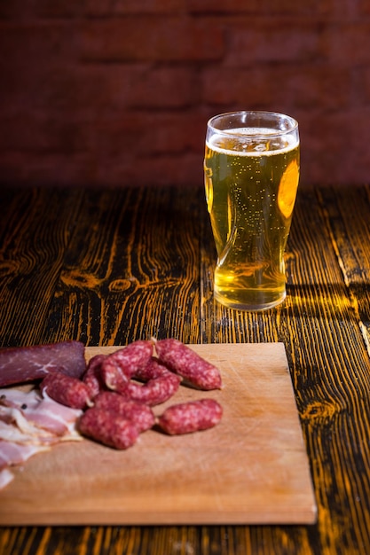 Close up of cutting board with sausages, bacon and meat on wooden table near a full glass of beer