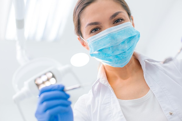 Close up of a dentist in a medical mask leaning towards her patient with a tool in hand