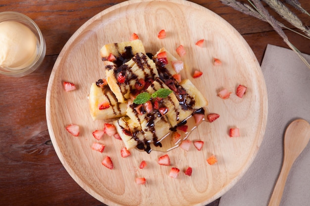 Close up of dessert which has Bread, strawberry berries, caramel and put peppermint leaf on top on wood plate and on the wood table