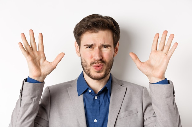 Close-up of disappointed and disgusted businessman raising hands up, grimacing from dislike and aversion, standing reluctant on white background.